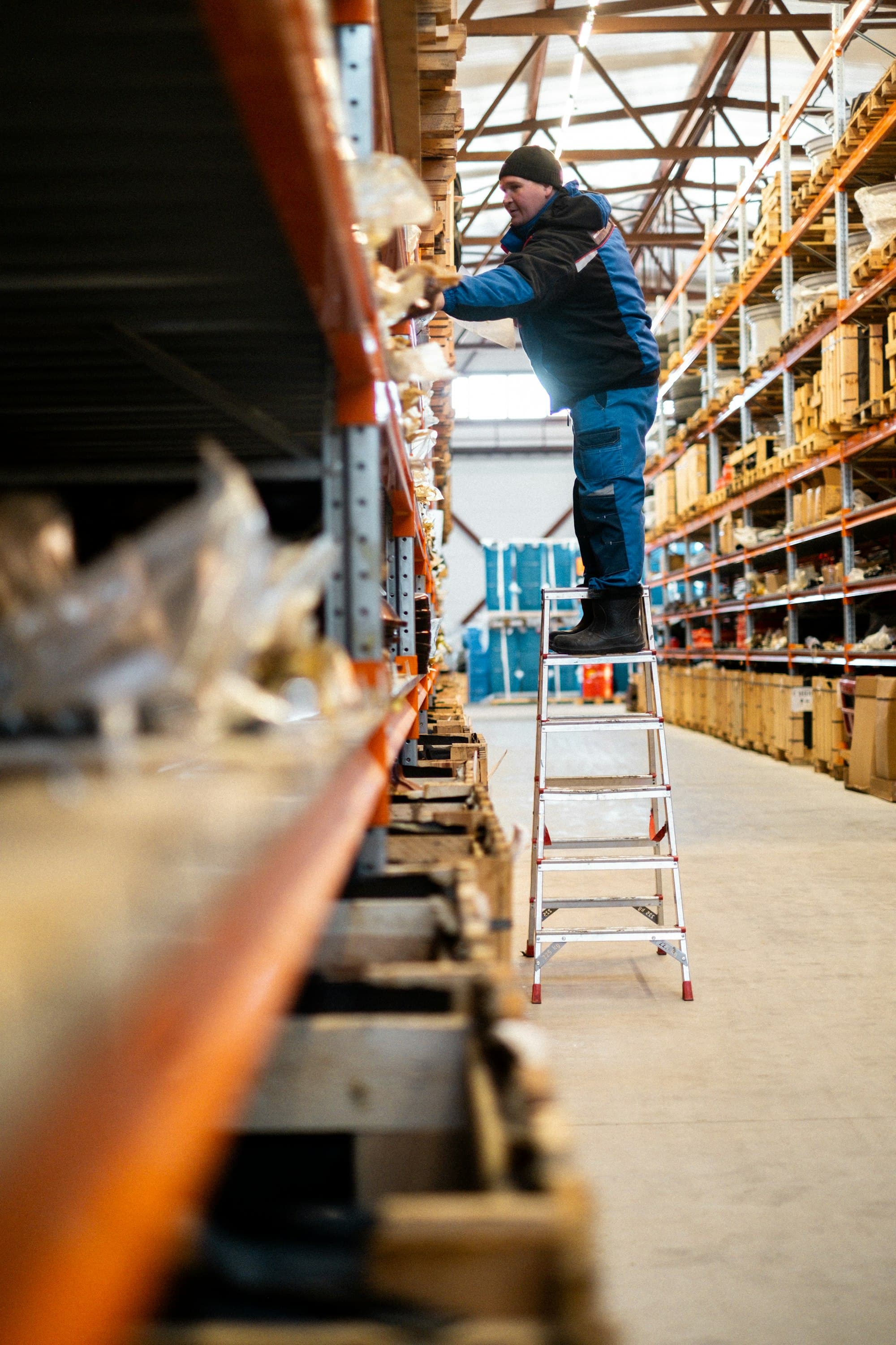 Warehouse worker in blue overalls reaching for inventory on a ladder